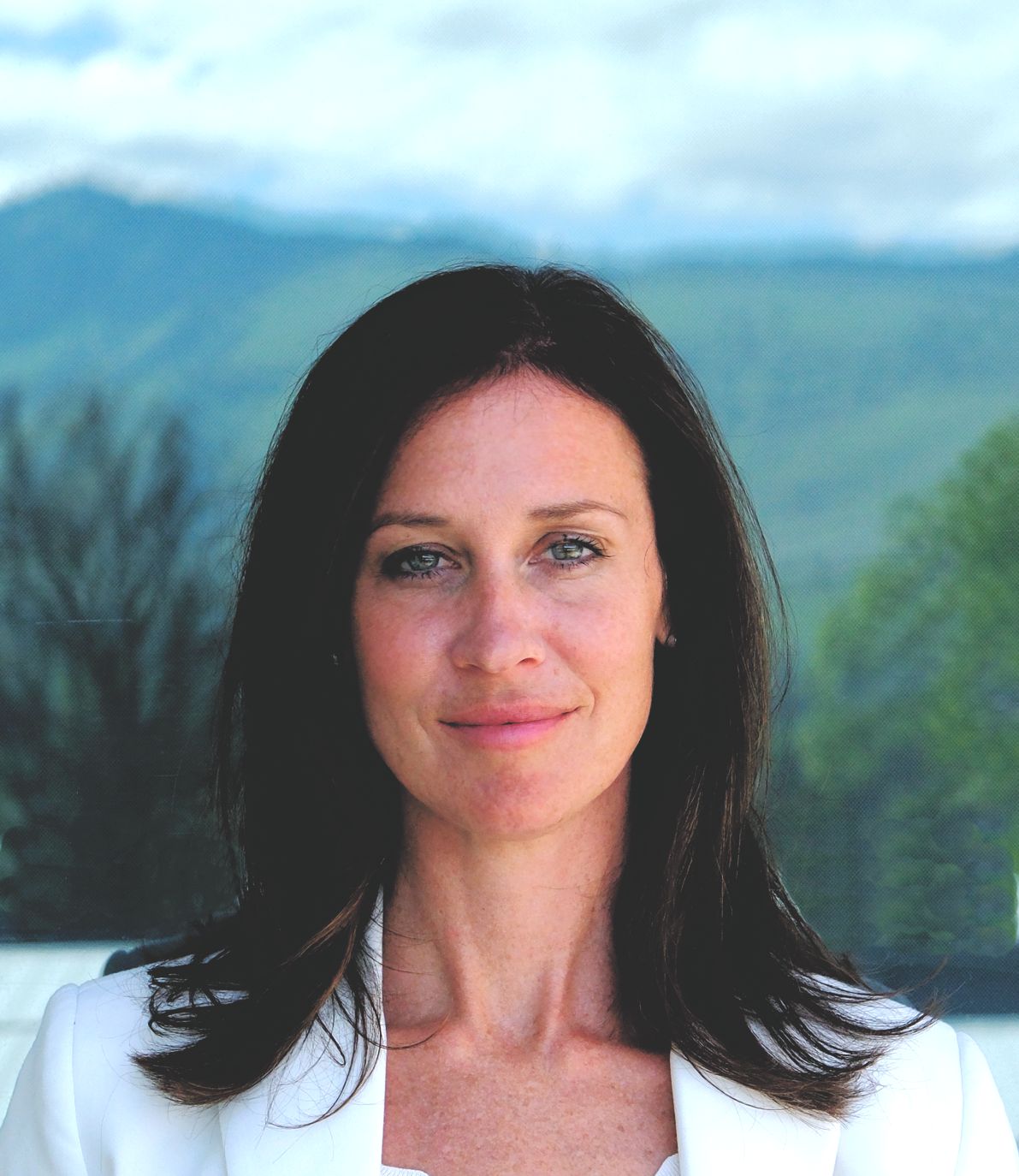 Woman with shoulder-length dark hair wearing a white jacket with a mountain and greenery background.