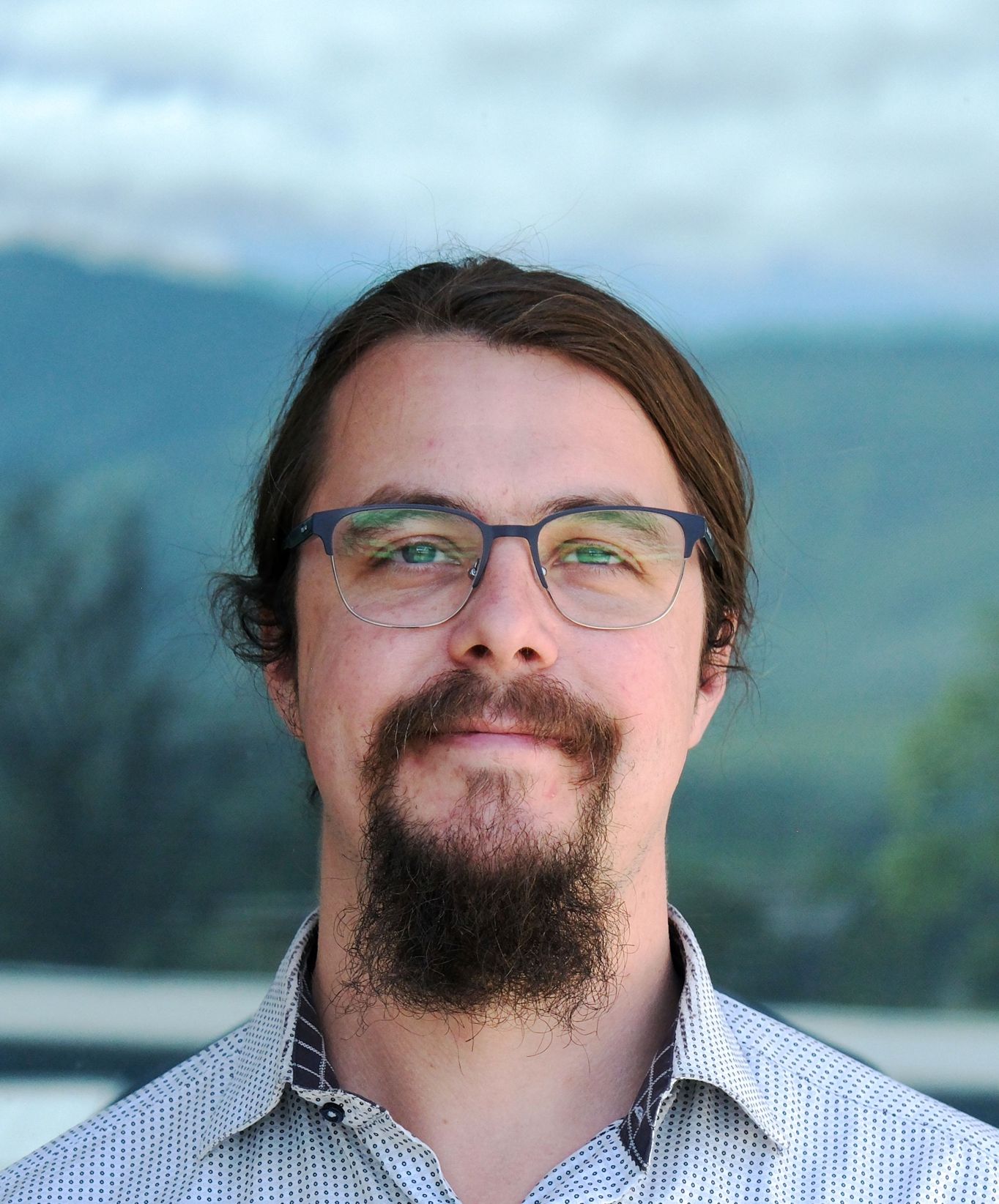 Man with glasses, long dark hair tied back, and a goatee smiling slightly against a blurred outdoor background.
