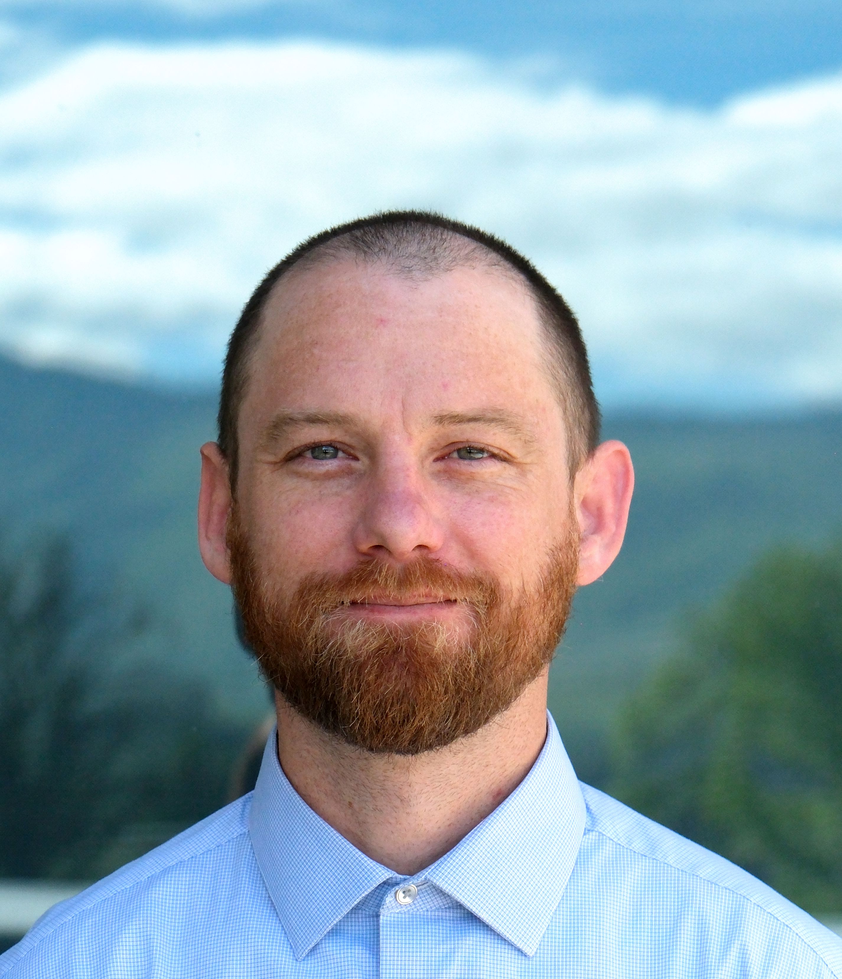 Smiling man with short hair and ginger beard wearing a light blue checkered shirt with a buttoned collar outdoors.