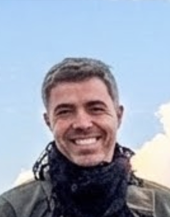 Smiling man with short gray hair wearing a dark scarf and jacket against a blue sky background.